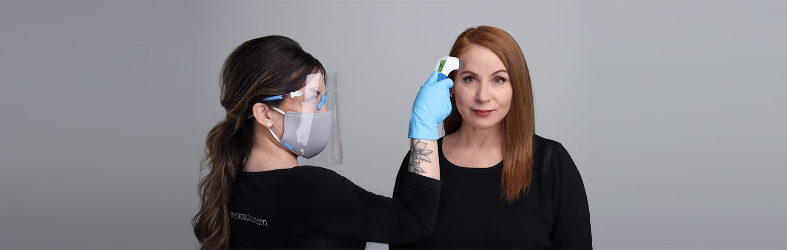 Image of Esthetician Wearing a mask and using a Thermometor on a client with red hair who is wearing a black shit.