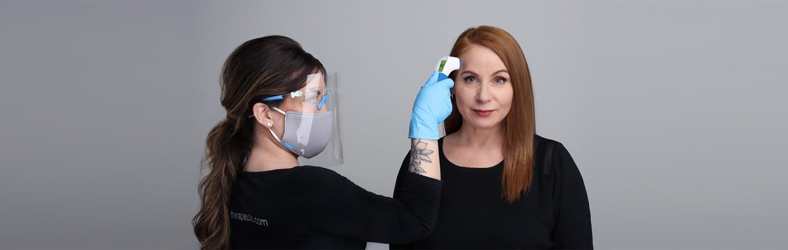 Image of Esthetician Wearing a mask and using a Thermometor on a client with red hair who is wearing a black shit. 